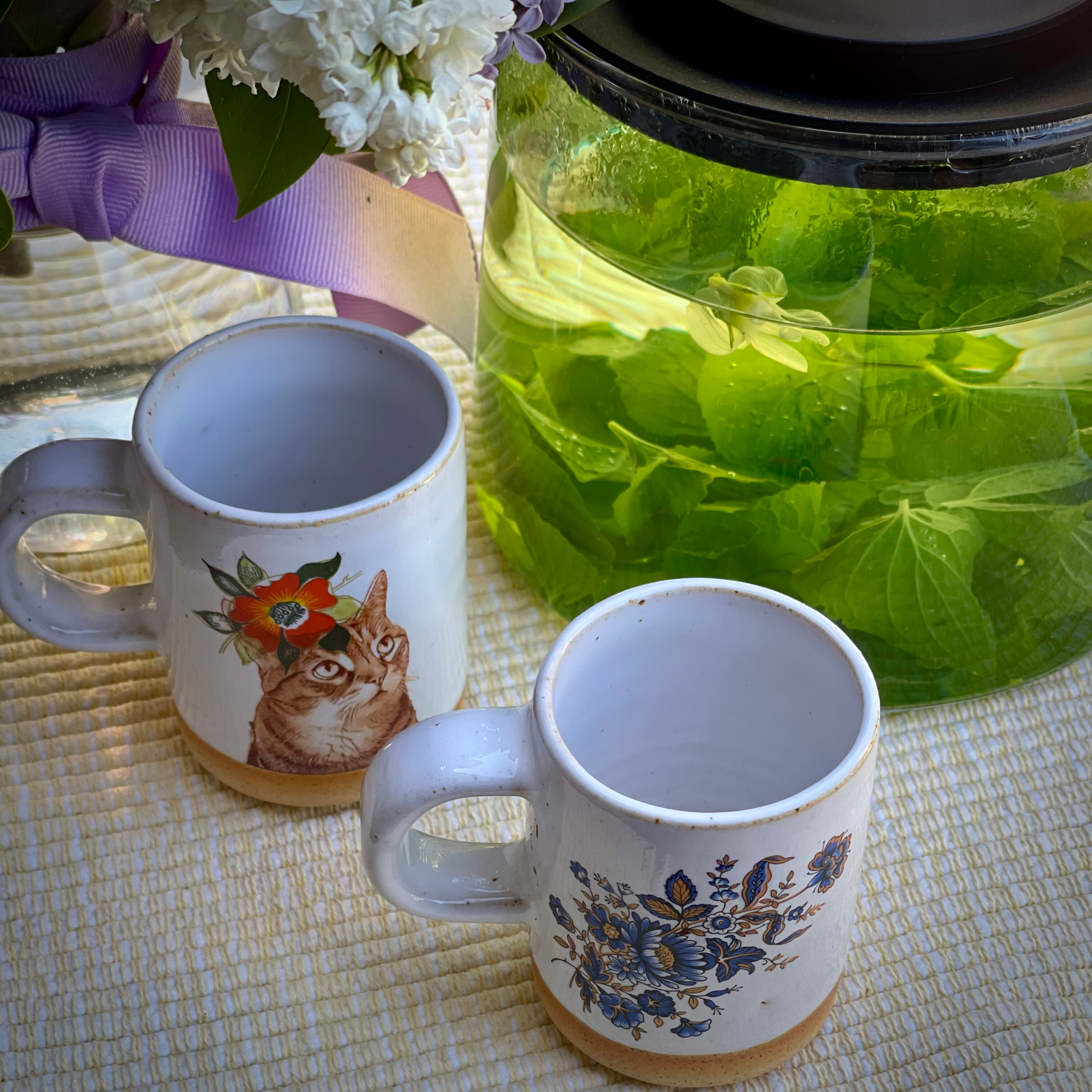 Two ceramic cups beside a large glass jar of green herbal tea, with flowers nearby on a table.